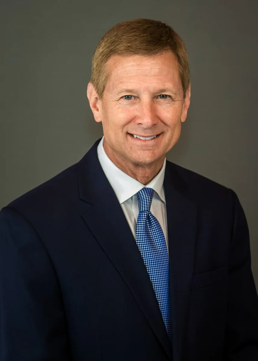 Professional corporate headshot of a man in a dark suit and blue tie against a gray background