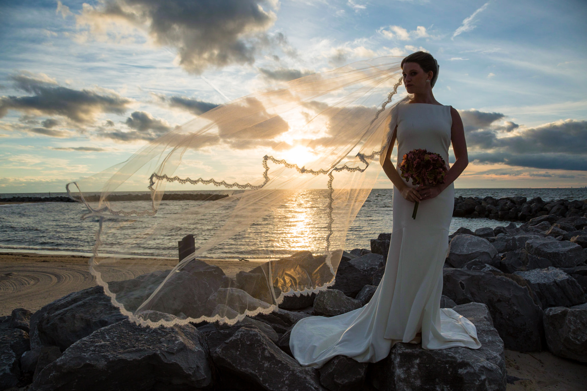 A bride standing on beach rocks at sunset with her veil blowing in the wind